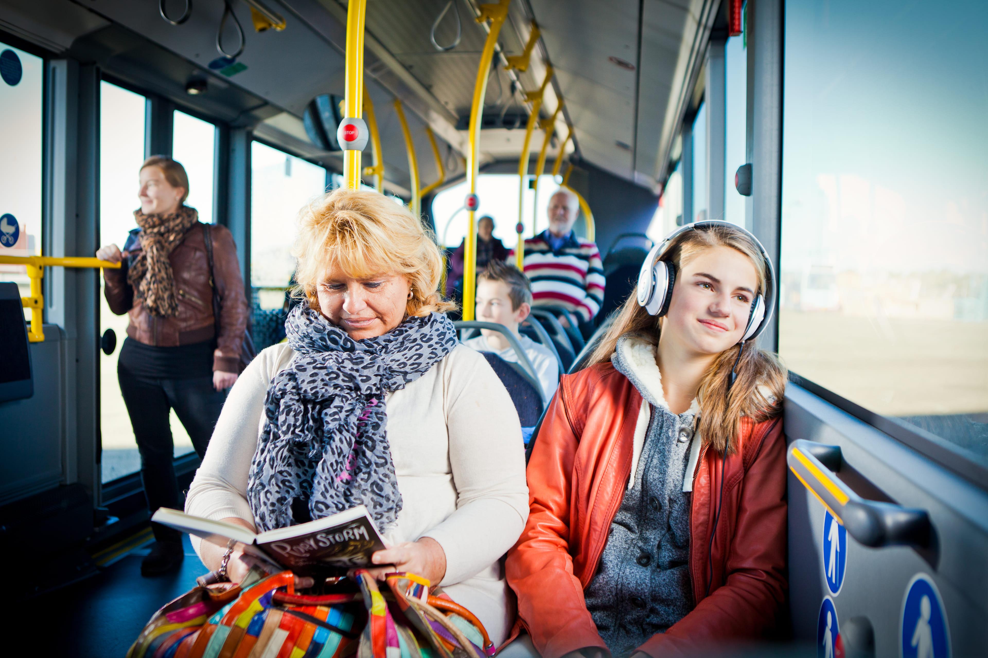 Twee vrouwen zitten naast elkaar in de bus. Een leest een tijdschrift de ander kijkt uit het raam met een koptelefoon.